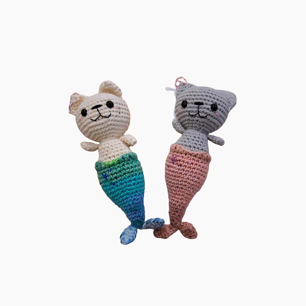 Two crocheted mermaid cat toys on a plain light background. One toy is cream-colored with a blue and green gradient mermaid tail, and the other is gray with a pink mermaid tail.