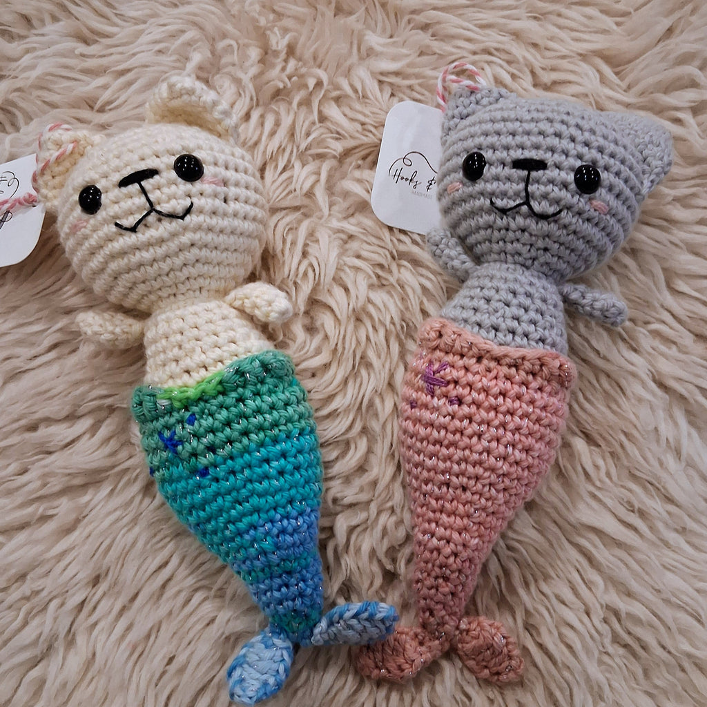 Two crocheted mermaid toys, one white and one gray, on a fluffy beige surface.