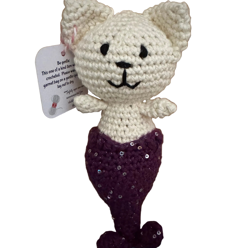 Crocheted mermaid cat-shaped toy with a purple sequin tail on a wooden surface.