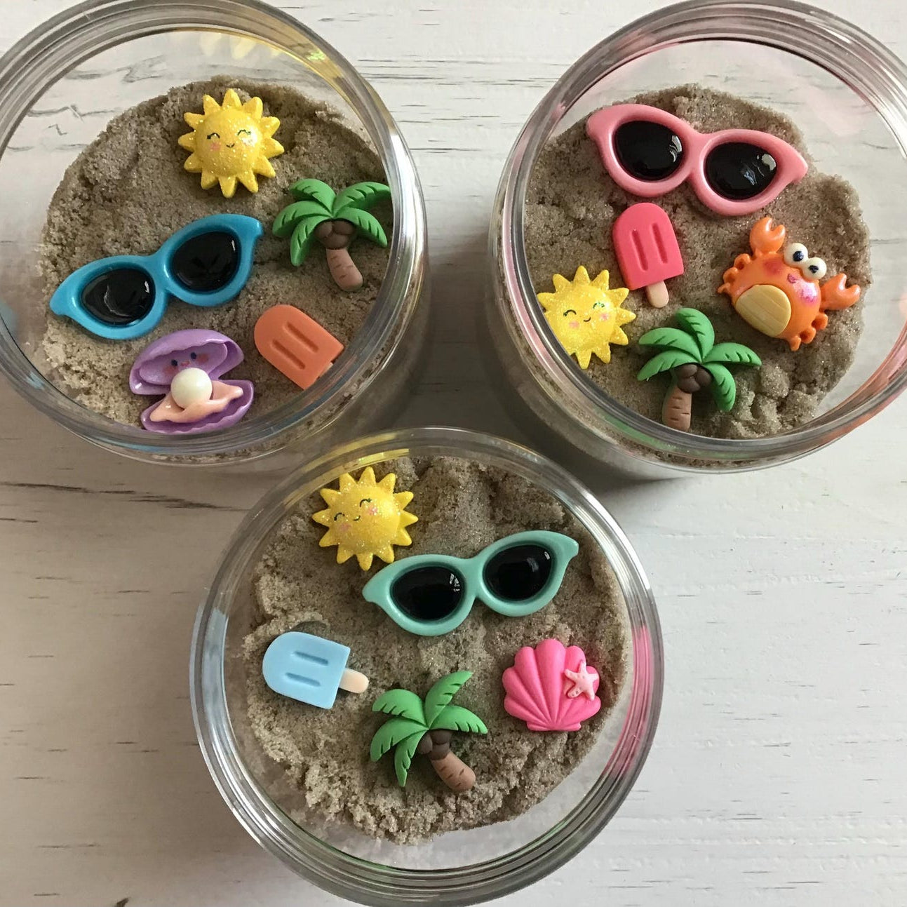 Three small glass jars filled with sand and decorated with colorful beach-themed figurines on a light wooden surface.
