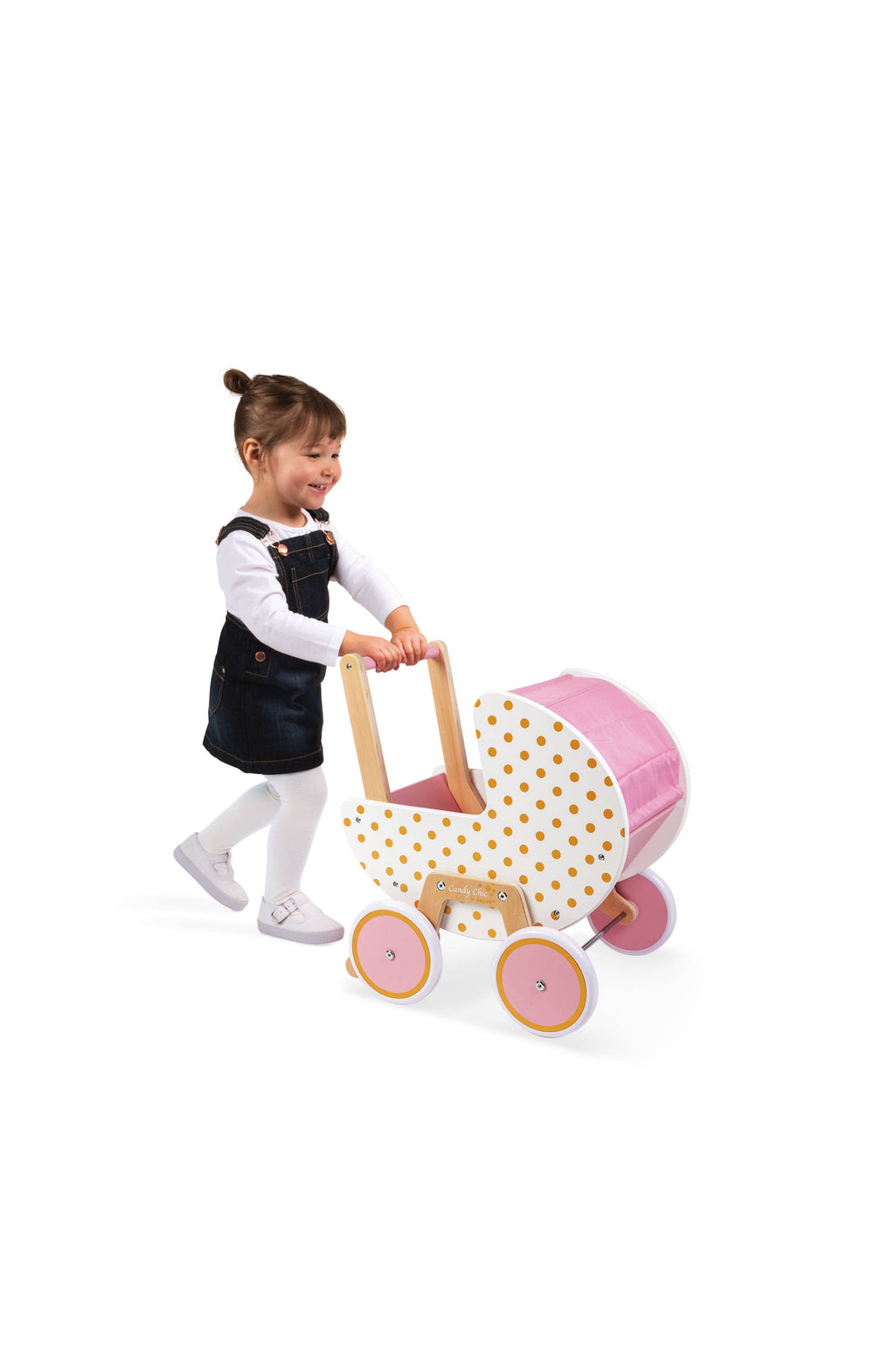 A young child wearing a navy denim pinafore dress over a white long-sleeve shirt pushes a wooden toy baby stroller with pink fabric canopy and polka-dot pattern, set against a solid white background.