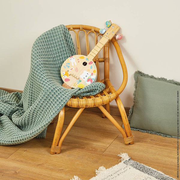 Wicker chair with a green textured blanket and a decorative ukulele on a wooden floor.