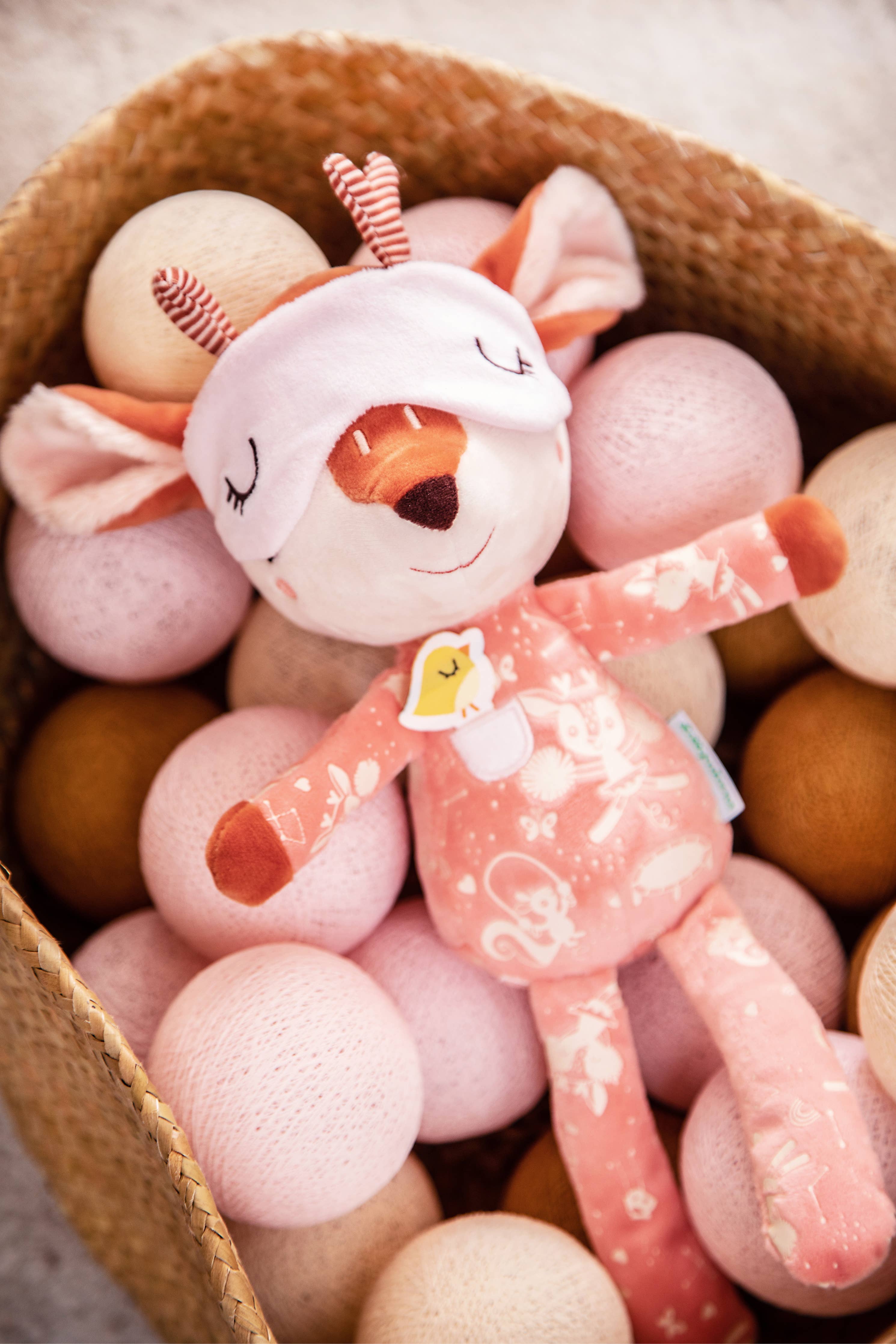 Plush toy deer in pajamas surrounded by pink and orange balls in a woven basket.