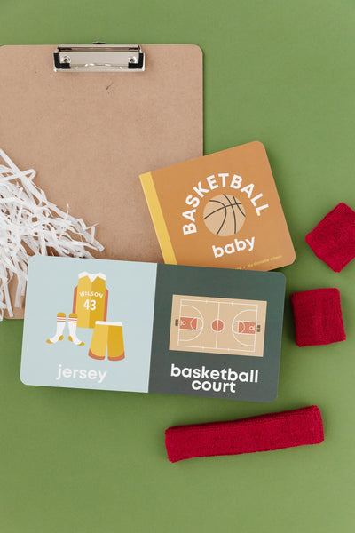Basketball-themed baby items including a book, cards, and headbands on a green background