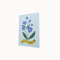 A vertical greeting card featuring a blue forget-me-not flower illustration with green leaves and a yellow banner that reads 'THINKING OF YOU', shown against a plain white background.