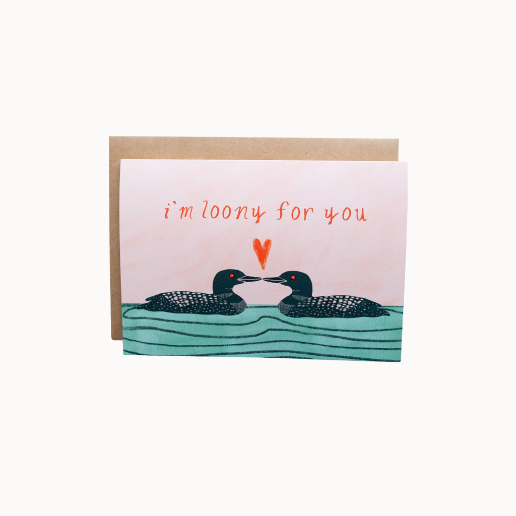 A square greeting card with a kraft envelope, featuring an illustration of two loons facing each other above wavy teal water, with the phrase 'i'm loony for you' in orange handwritten text and a small orange heart between them.