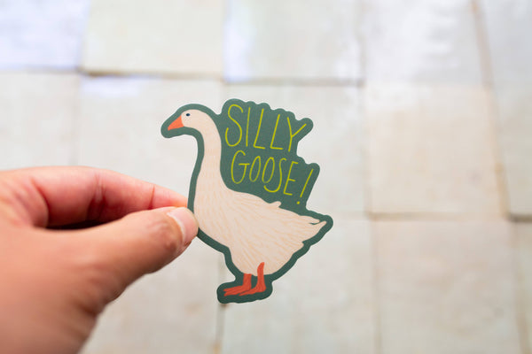 Hand holding a sticker of a goose with 'Silly Goose!' text against a tiled wall background
