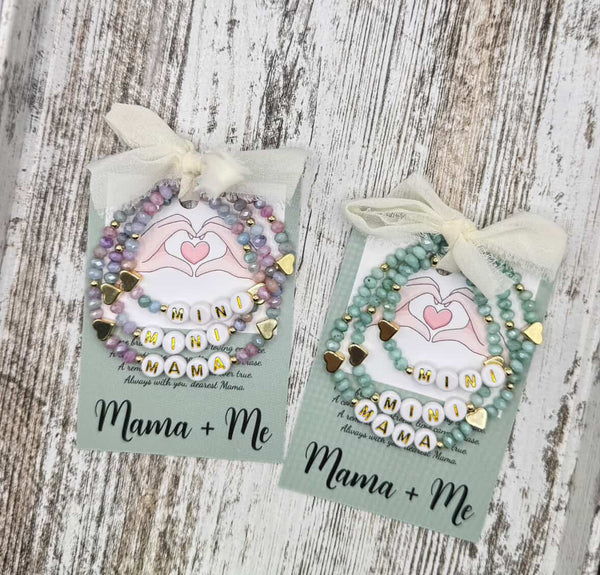 Two 'Mama + Me' beaded bracelets on cards with ribbons against a wooden background.