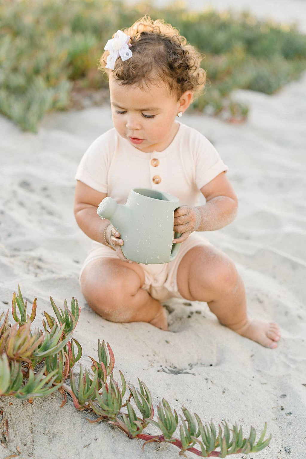 Child sitting on a sandy beach holding a green watering can