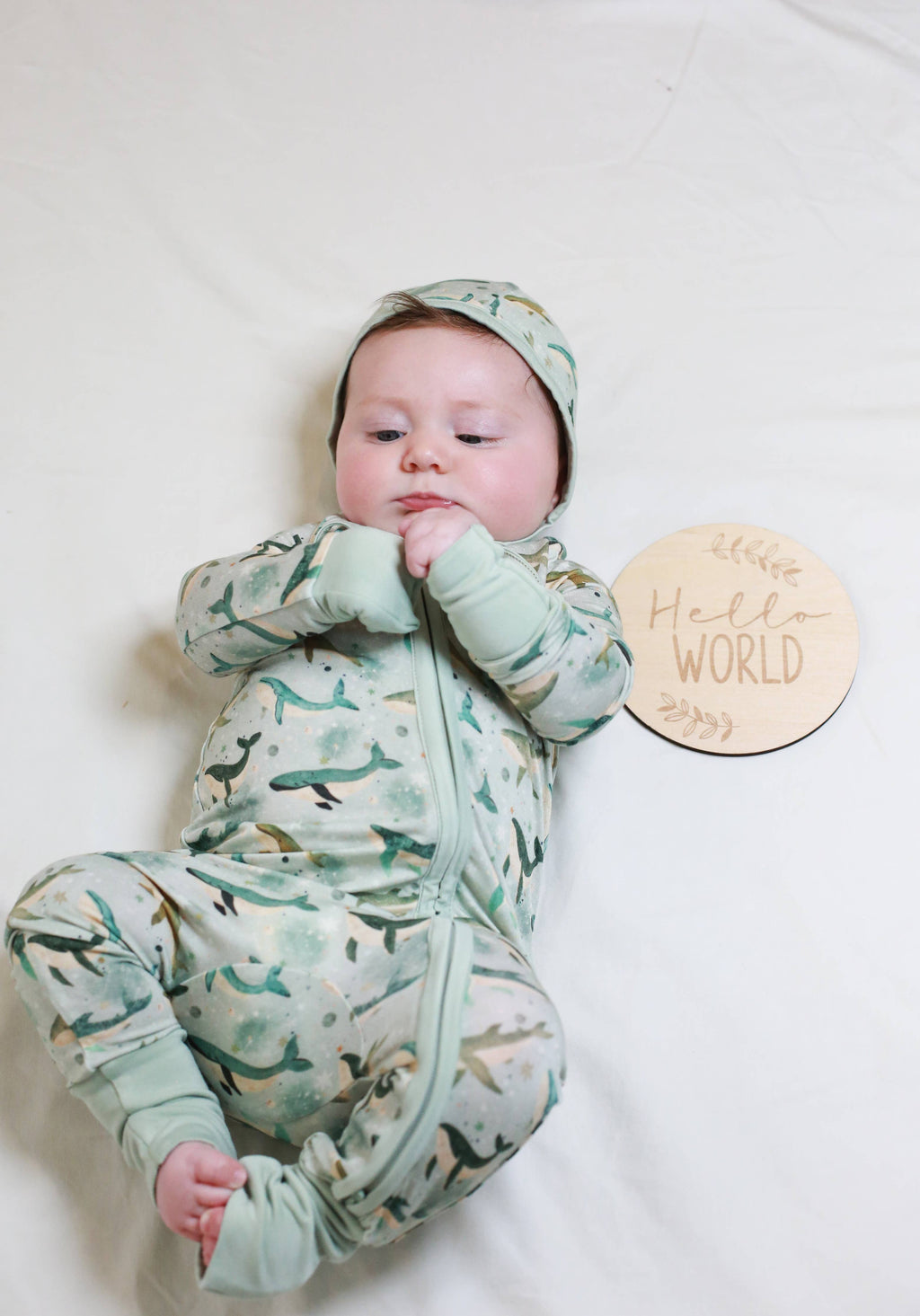 A baby lying on a white blanket wearing a sage green and white dinosaur print footed onesie and matching hat. A round wooden sign that reads 'Hello WORLD' is placed next to the baby.