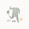 Baby one-piece pajama set laid flat on a white background, including a long-sleeve zip pajama with a dinosaur print, a matching bonnet, a headband, and a small box. The pajama and accessories are pale mint green with a multicolor dinosaur pattern.