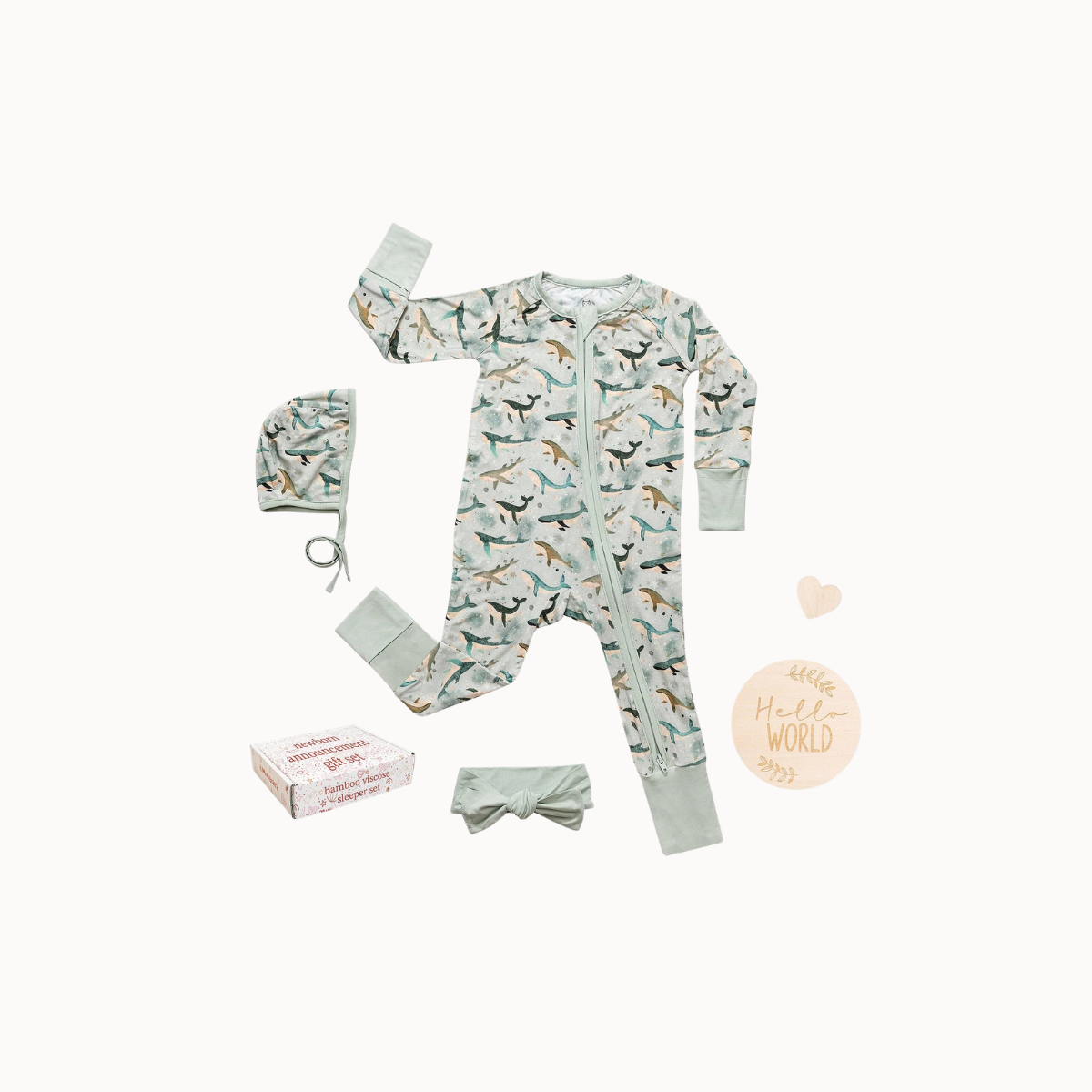 Baby one-piece pajama set laid flat on a white background, including a long-sleeve zip pajama with a dinosaur print, a matching bonnet, a headband, and a small box. The pajama and accessories are pale mint green with a multicolor dinosaur pattern.