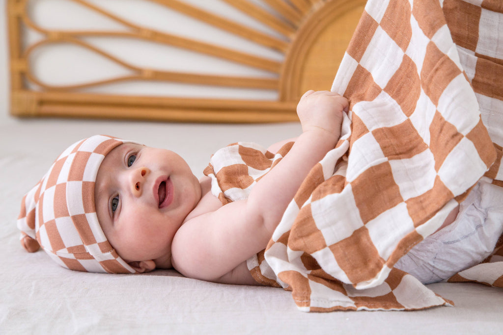 Baby wrapped in a checkered blanket and headband, lying on a light surface with a decorative wall in the background.