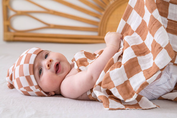 Baby wrapped in a checkered blanket and headband, lying on a light surface with a decorative wall in the background.