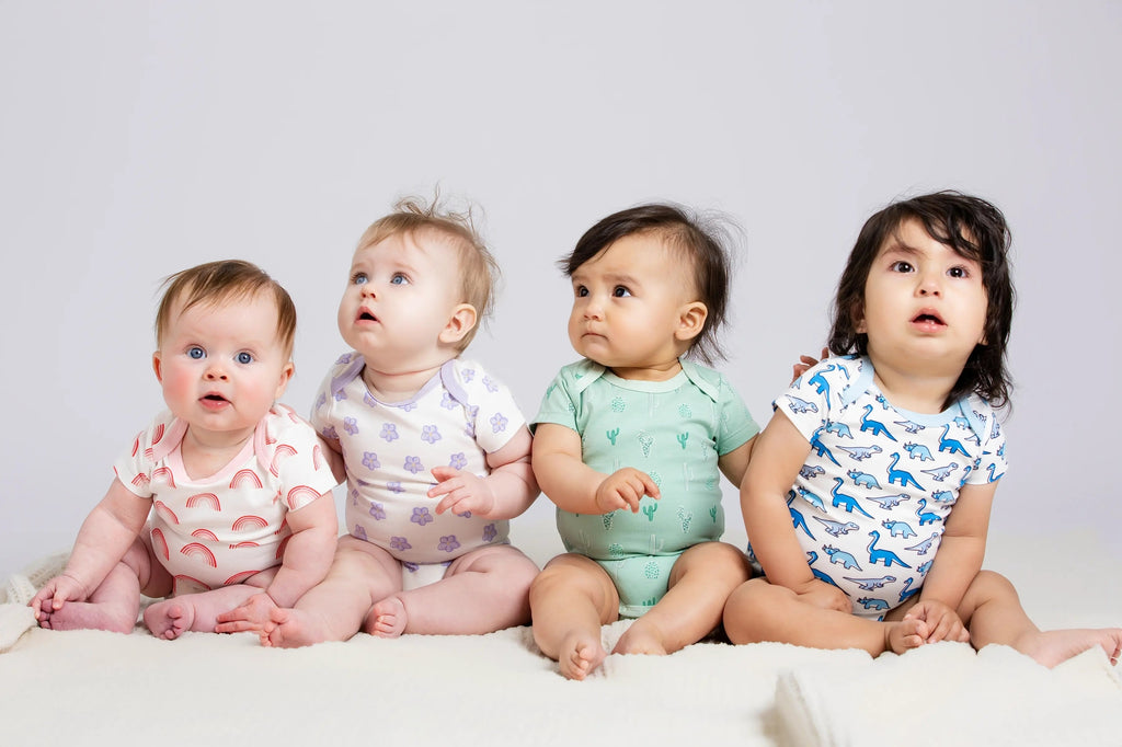 Four babies sitting on a white blanket wearing colorful outfits against a light gray background