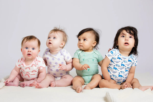 Four babies sitting on a white blanket wearing colorful outfits against a light gray background