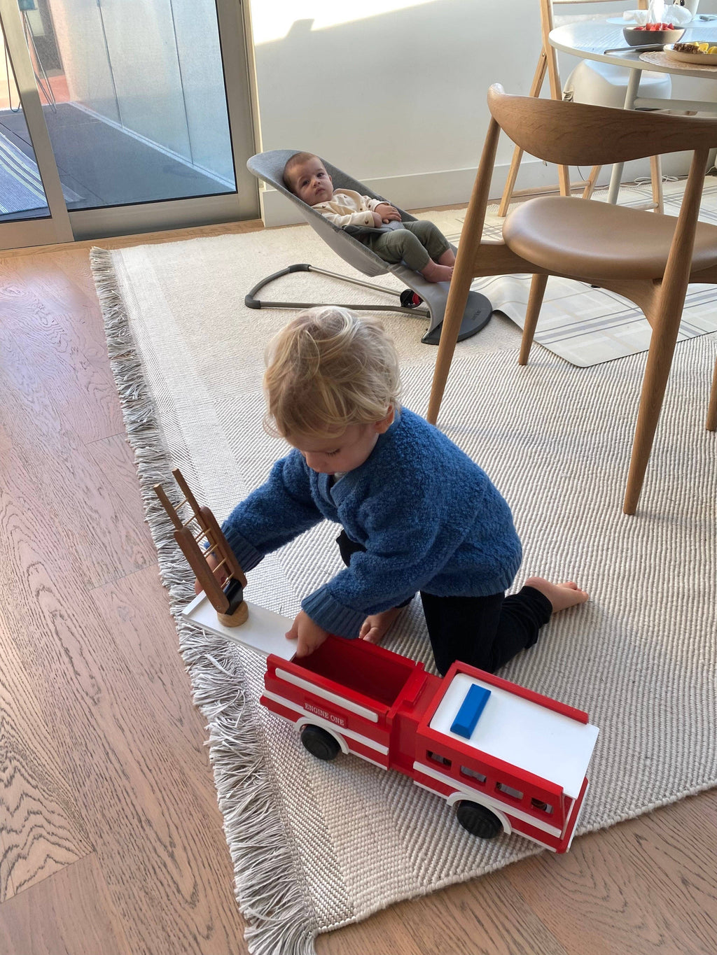 A toddler in a blue sweater kneels on a beige rug, playing with a red and white wooden toy fire truck. In the background, another child sits in a gray baby chair in a bright, modern room with wooden floors and dining chairs.