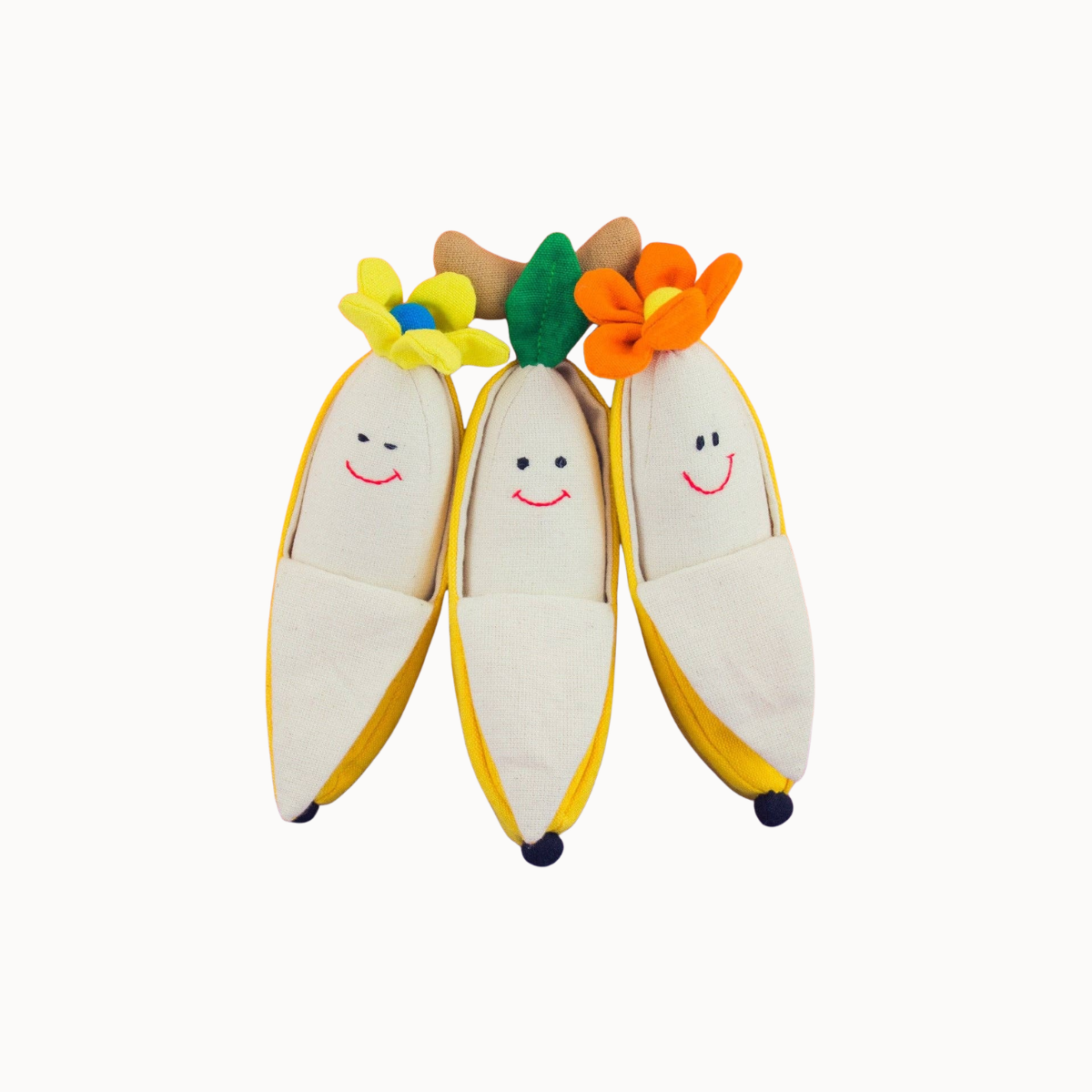 Three plush banana-shaped toys with embroidered smiling faces and colorful flower tops, arranged side by side on a plain light background.
