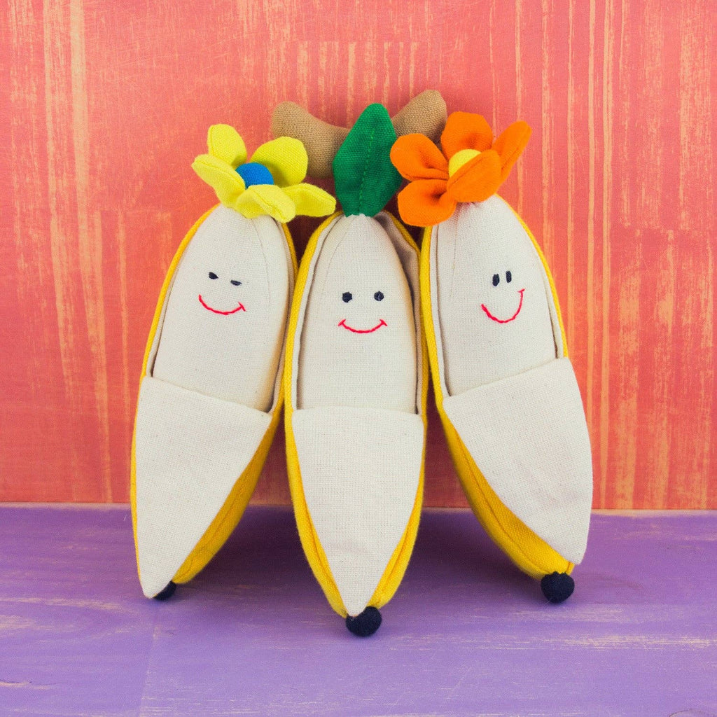 Three plush banana-shaped slippers with embroidered smiling faces, displayed on a purple surface against a textured orange-pink background. The slippers have cream-colored bodies, yellow peel edges, and colorful flower crowns in yellow, green, and orange.