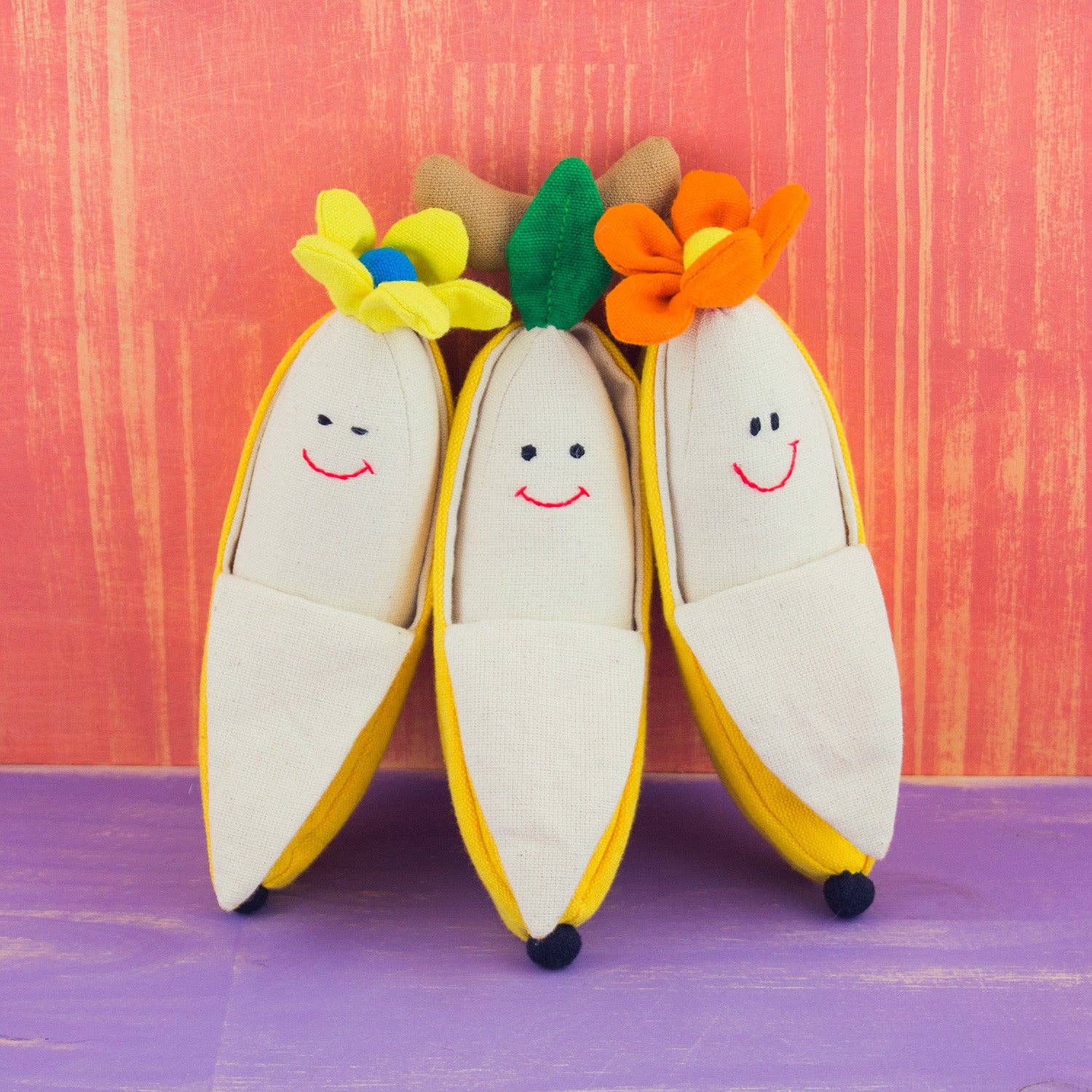 Three plush banana-shaped slippers with embroidered smiling faces, displayed on a purple surface against a textured orange-pink background. The slippers have cream-colored bodies, yellow peel edges, and colorful flower crowns in yellow, green, and orange.