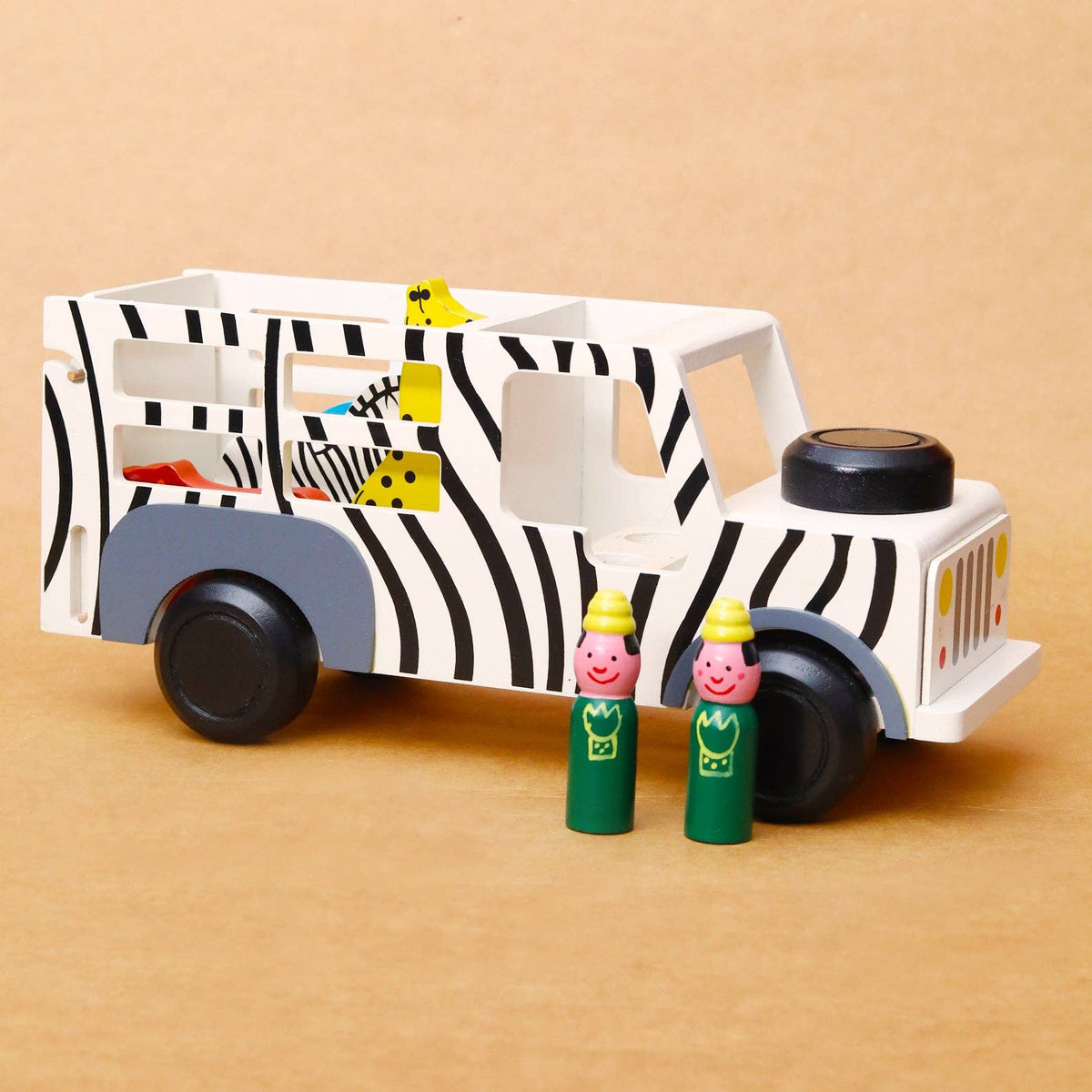 A wooden toy safari bus with black and white zebra stripes, gray wheels and fenders, and small animal and bird figures inside, shown on a warm tan background with two small green peg dolls in front.