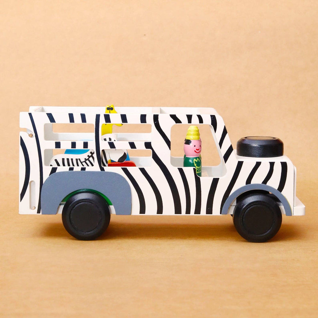 A wooden toy truck designed like a zebra bus, featuring black and white zebra stripes, gray wheel arches, and small colorful toy passengers inside, set against a solid tan background.