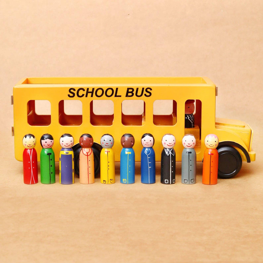 A yellow wooden toy school bus with the words 'SCHOOL BUS' printed on the side, shown with ten small painted peg children standing in front of it on a plain beige background.