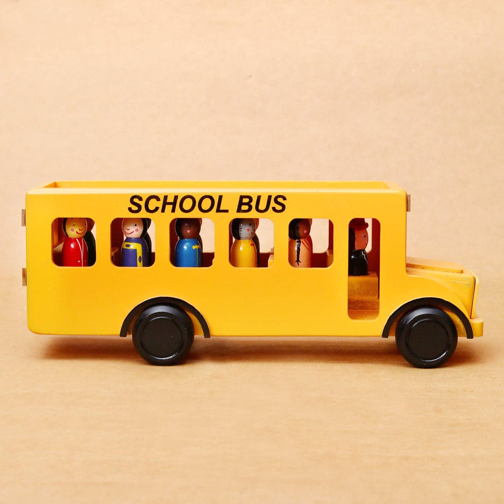 A yellow wooden toy school bus with black wheels and the words 'SCHOOL BUS' printed on the side, shown on a plain beige background. Small multicolored toy passengers are visible inside the windows.