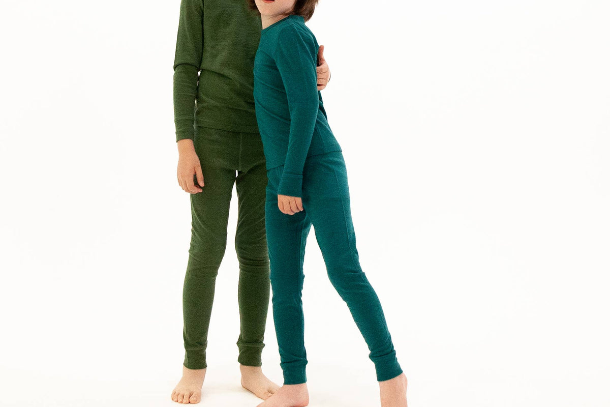 Two children modeling matching long-sleeve pajama sets, one in olive green and the other in teal, standing barefoot against a plain white background.