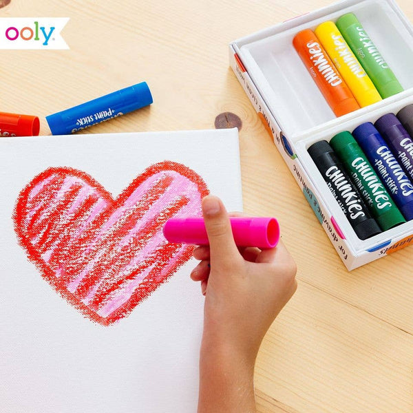 Child's hand drawing a heart with pink crayon on white paper, with a box of Crayola crayons in the background.