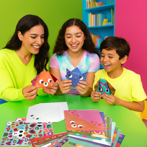 Three children and a woman sitting at a table with colorful paper crafts against a bright green wall.