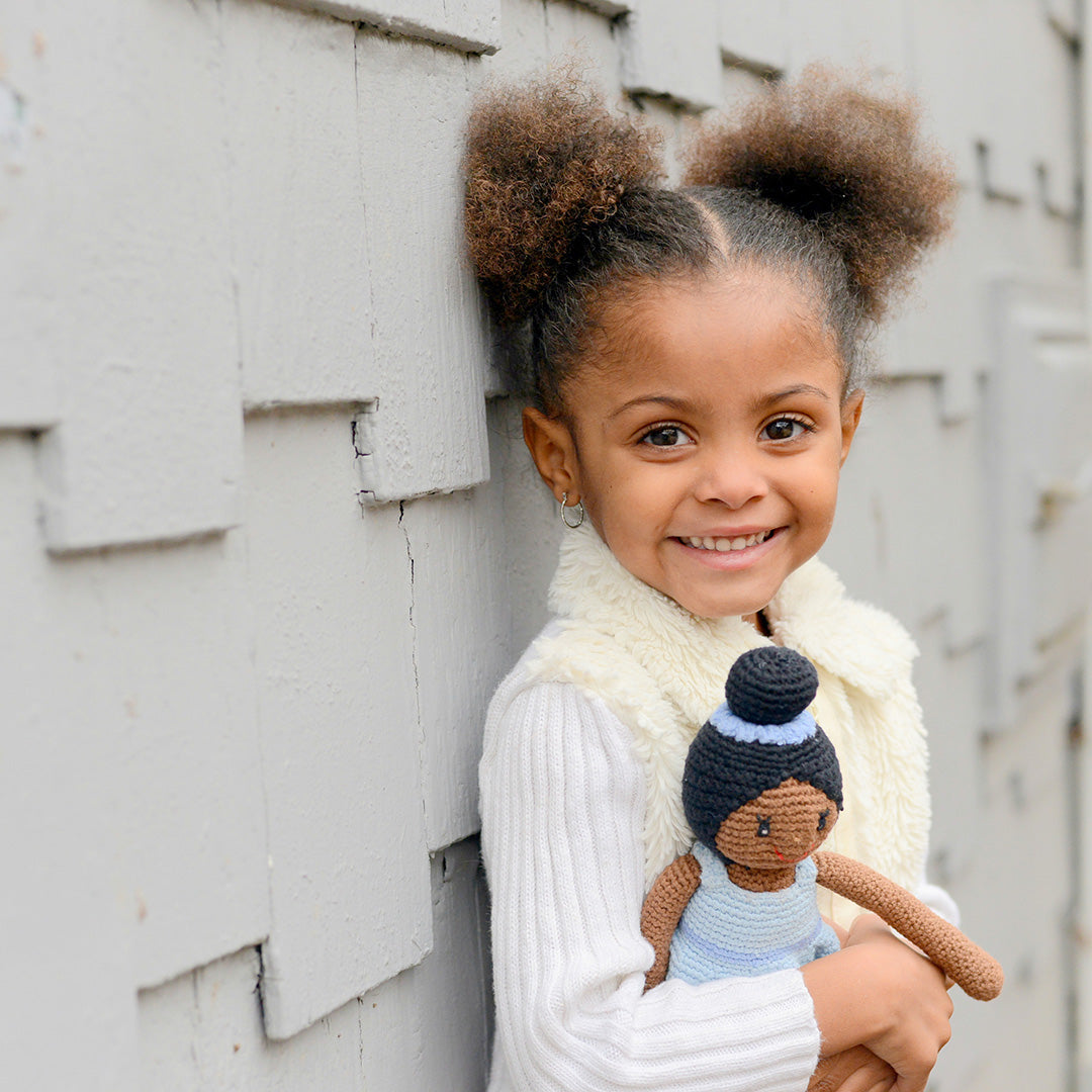 Young girl holding a small doll with a similar hairstyle against a wooden wall.