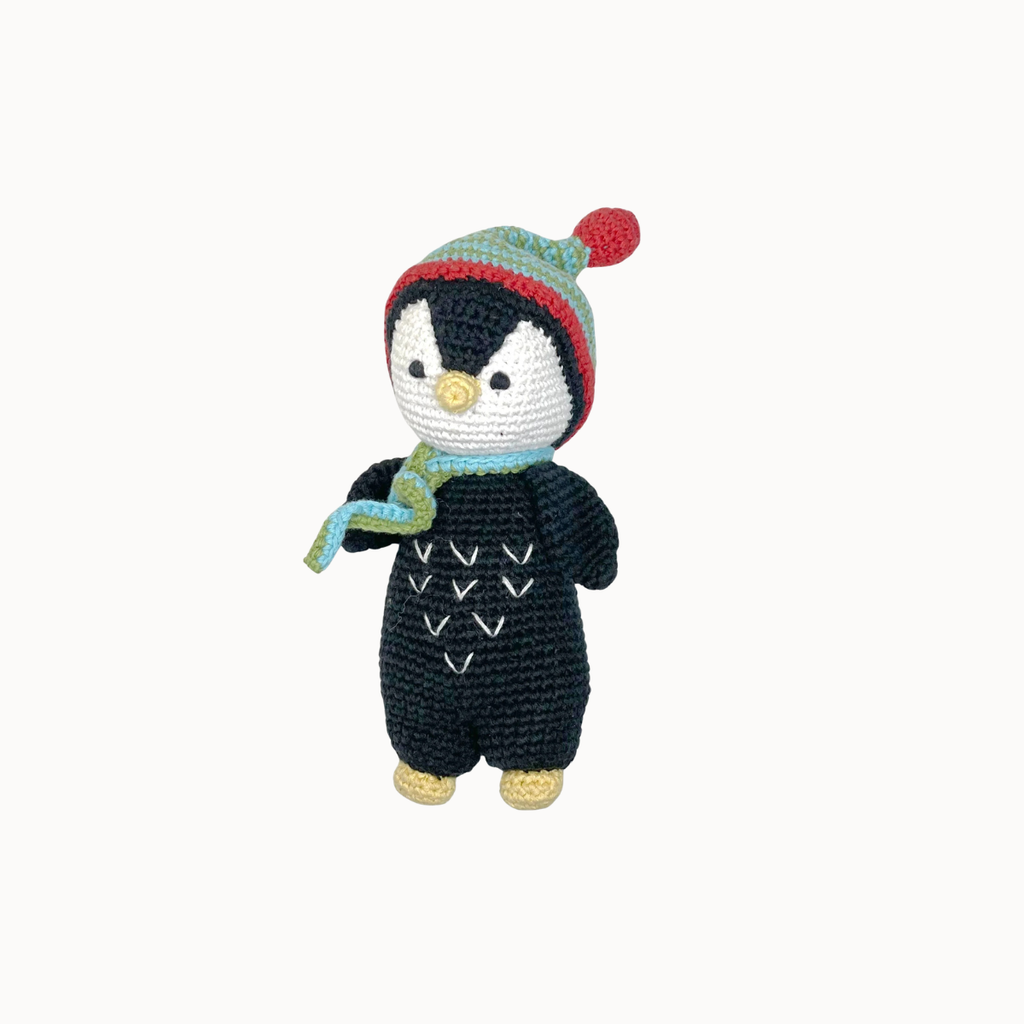 A crocheted penguin doll wearing a black onesie, a green and red striped knit hat with a red pom-pom, and a light blue scarf, set against a plain off-white background.