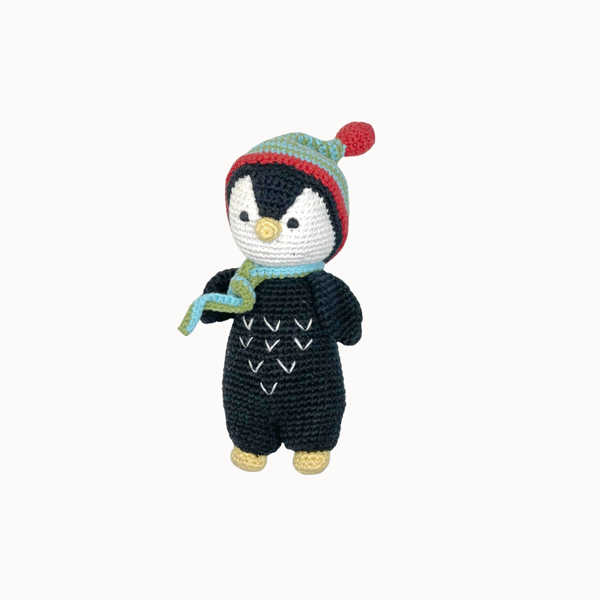 A crocheted penguin doll wearing a black onesie, a green and red striped knit hat with a red pom-pom, and a light blue scarf, set against a plain off-white background.