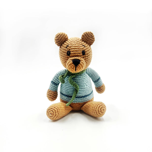 Crocheted teddy bear wearing a blue sweater with a green scarf on a white background