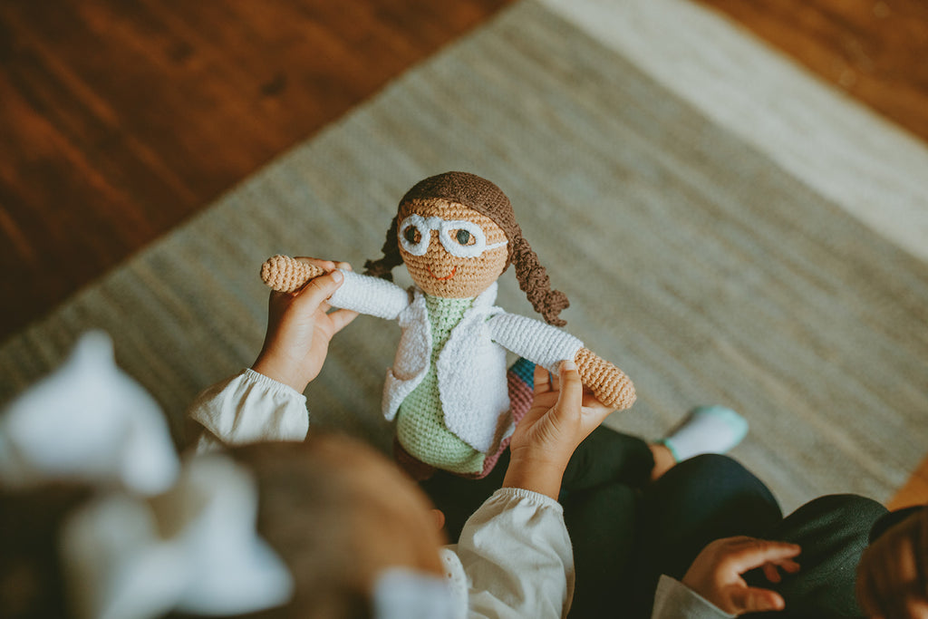 Person holding a knitted doll with glasses on a wooden floor.