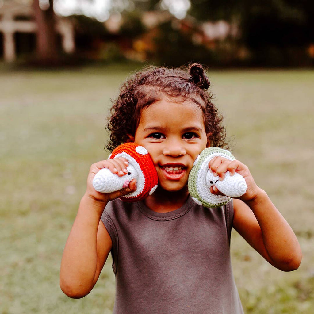A young child smiling outdoors while holding two crocheted plush hats, one red and white and one green and white, in front of their face.