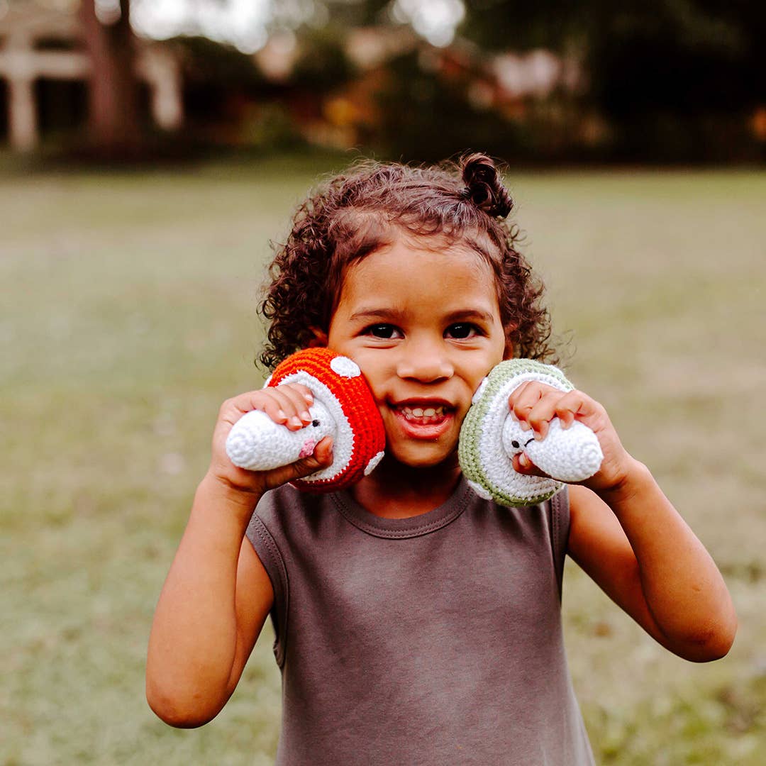 A young child smiling outdoors while holding two crocheted plush hats, one red and white and one green and white, in front of their face.