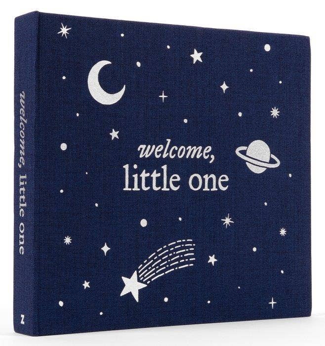 A square baby photo book with a navy blue linen-textured cover decorated with white celestial illustrations including stars, a crescent moon, a shooting star, and a planet. The front reads 'welcome, little one' in white script and serif text, and the spine repeats 'welcome, little one'.