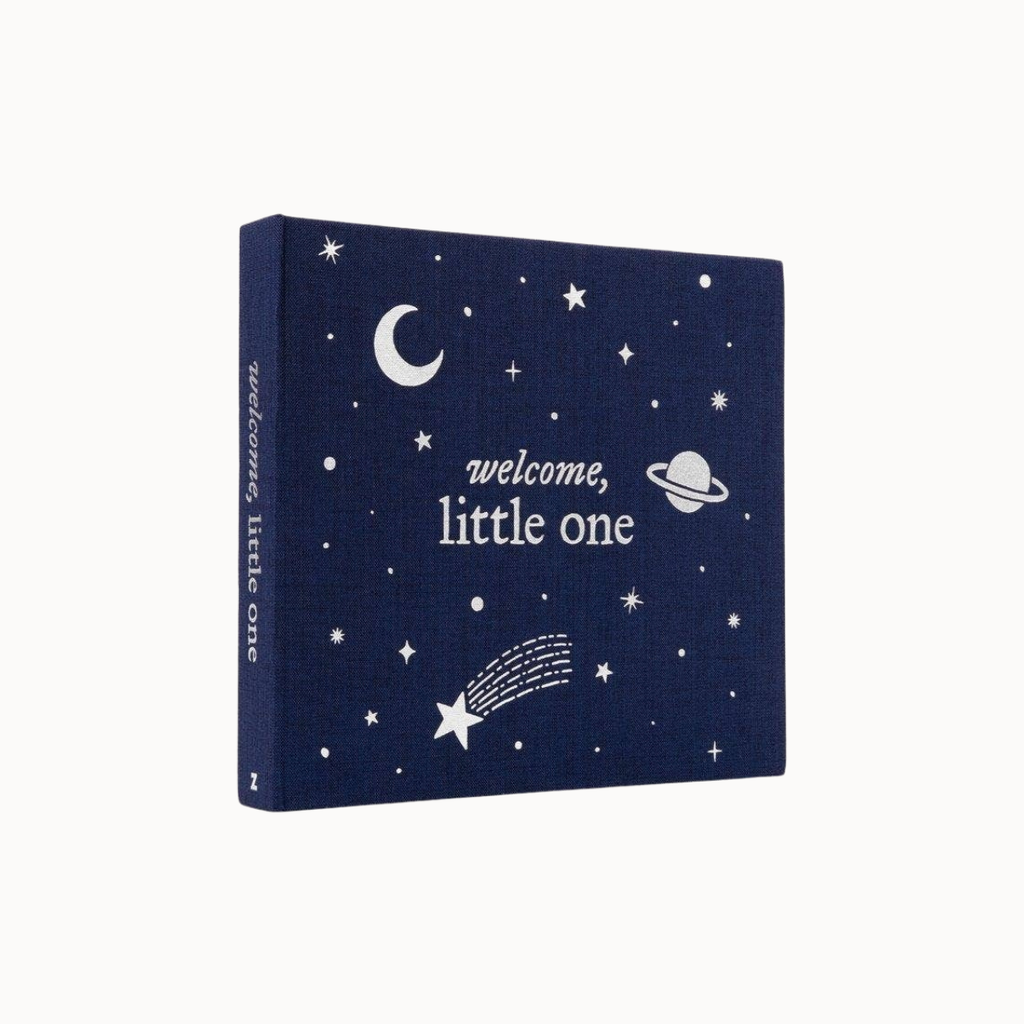 A square fabric baby keepsake box or book cover in dark navy blue, printed with white celestial illustrations including stars, a crescent moon, a planet, and a shooting star. The front reads 'welcome, little one' and the spine repeats the same text.