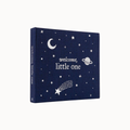 A square fabric baby keepsake box or book cover in dark navy blue, printed with white celestial illustrations including stars, a crescent moon, a planet, and a shooting star. The front reads 'welcome, little one' and the spine repeats the same text.