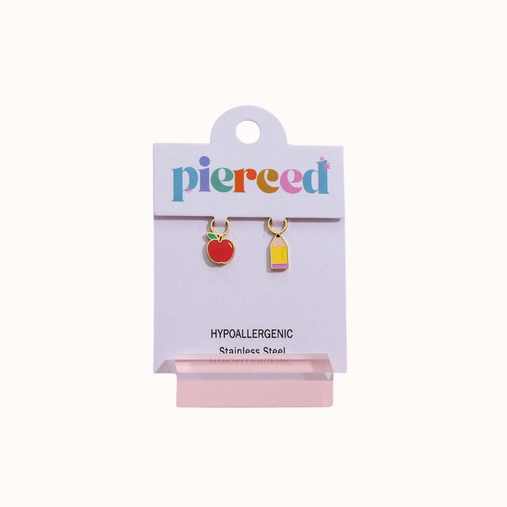 A pair of small hypoallergenic stainless steel earrings on a white display card. The left earring is shaped like a red apple with a green stem, and the right earring is a small yellow and pink rectangular charm.