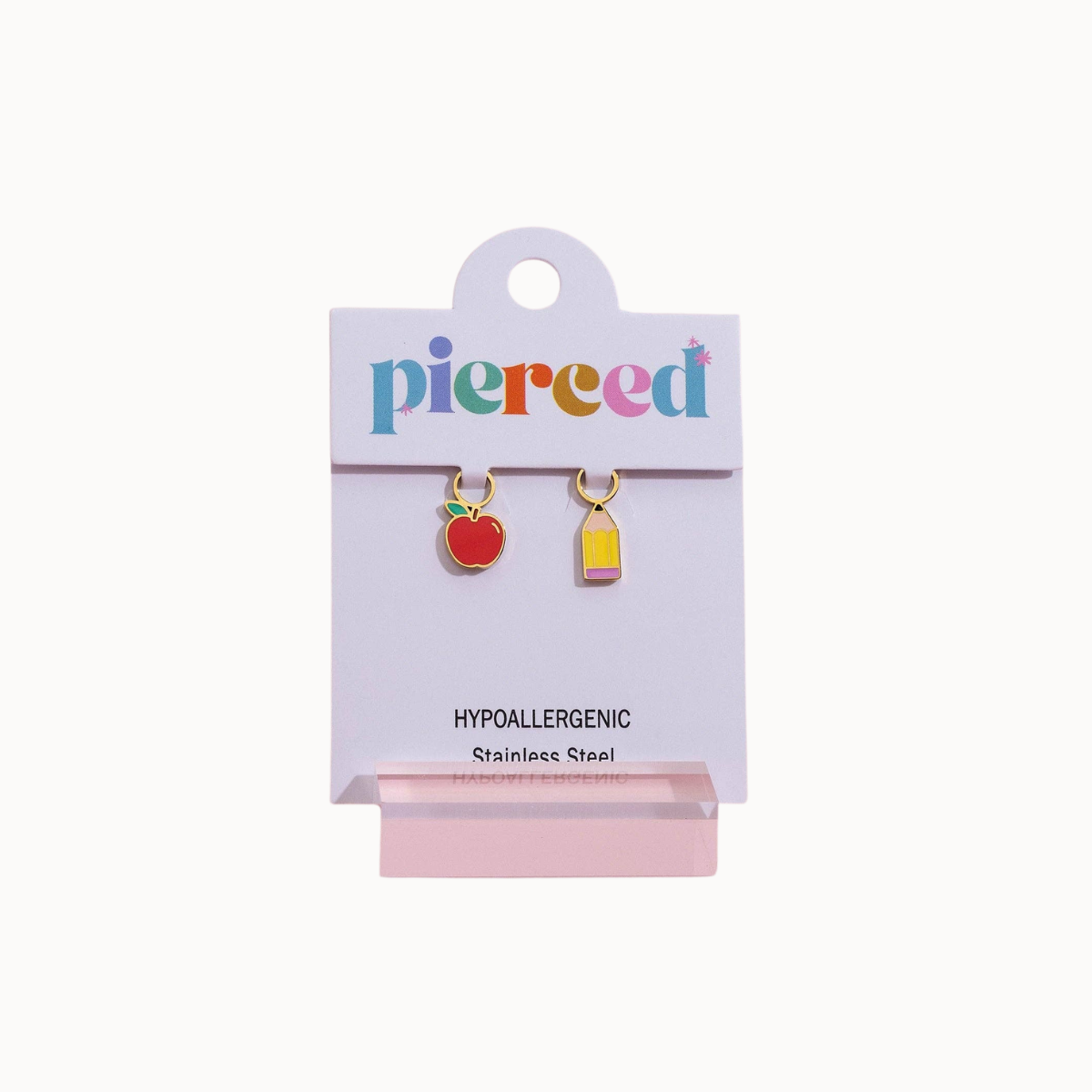 A pair of small hypoallergenic stainless steel earrings on a white display card. The left earring is shaped like a red apple with a green stem, and the right earring is a small yellow and pink rectangular charm.