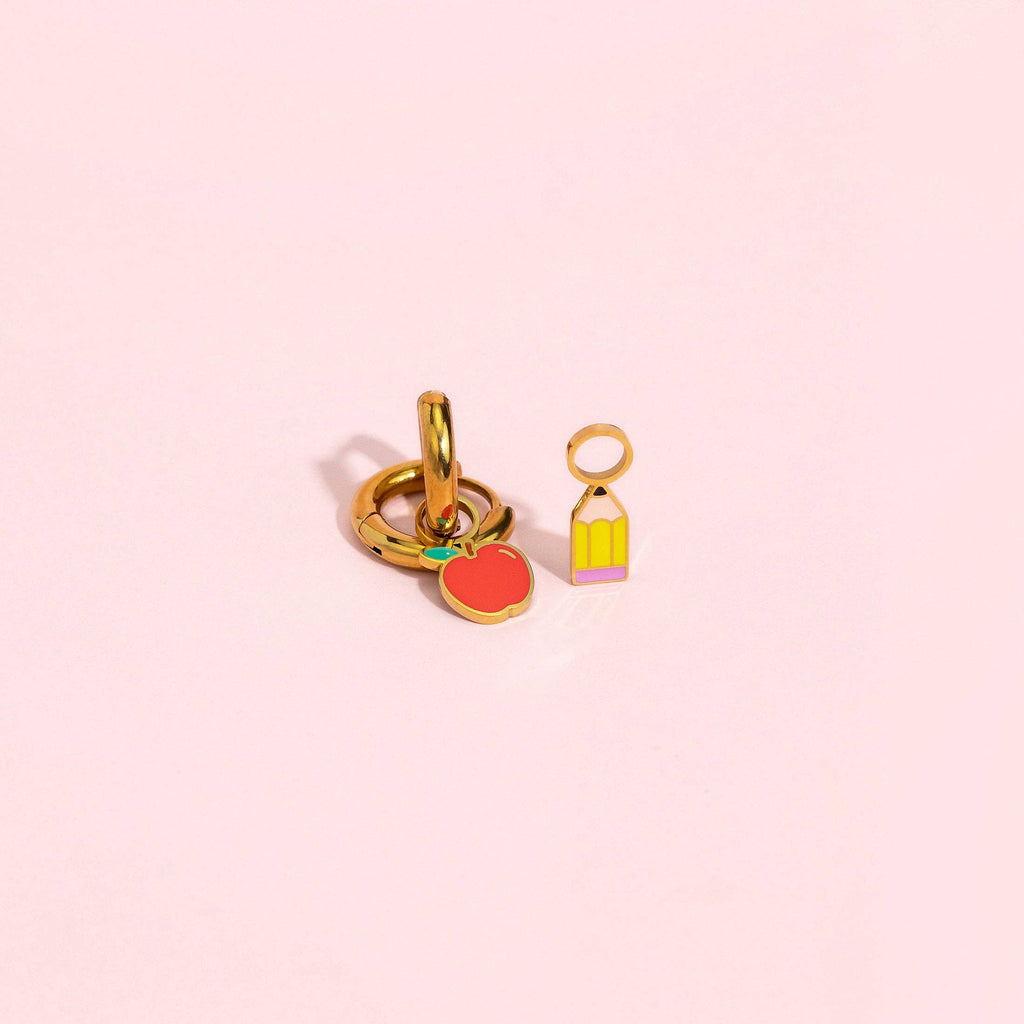 A pair of gold-plated charm links on a soft pink background. One charm is shaped like a red apple with a tiny green leaf, and the other is a small yellow birdcage with a pink base.