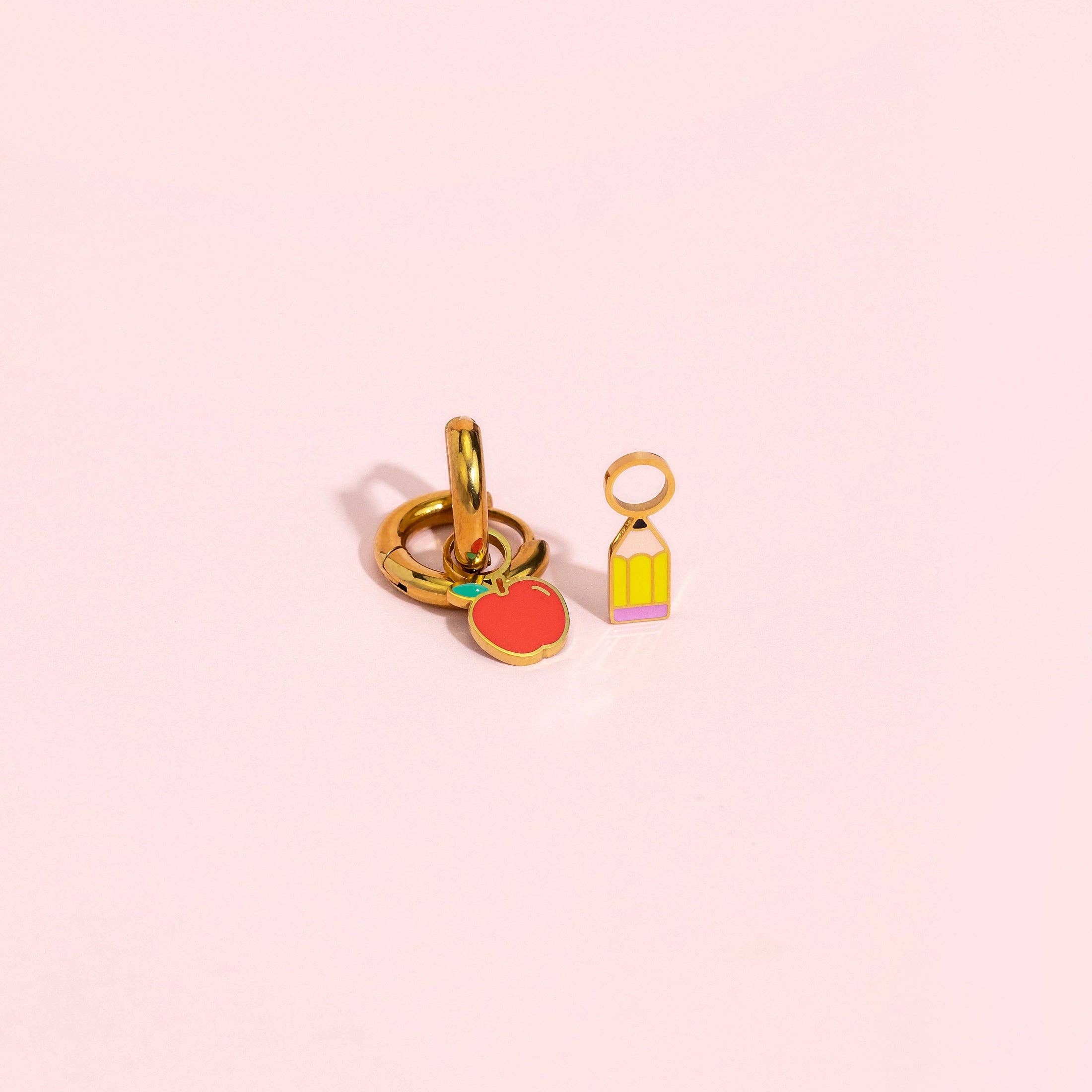 A pair of gold-plated charm links on a soft pink background. One charm is shaped like a red apple with a tiny green leaf, and the other is a small yellow birdcage with a pink base.