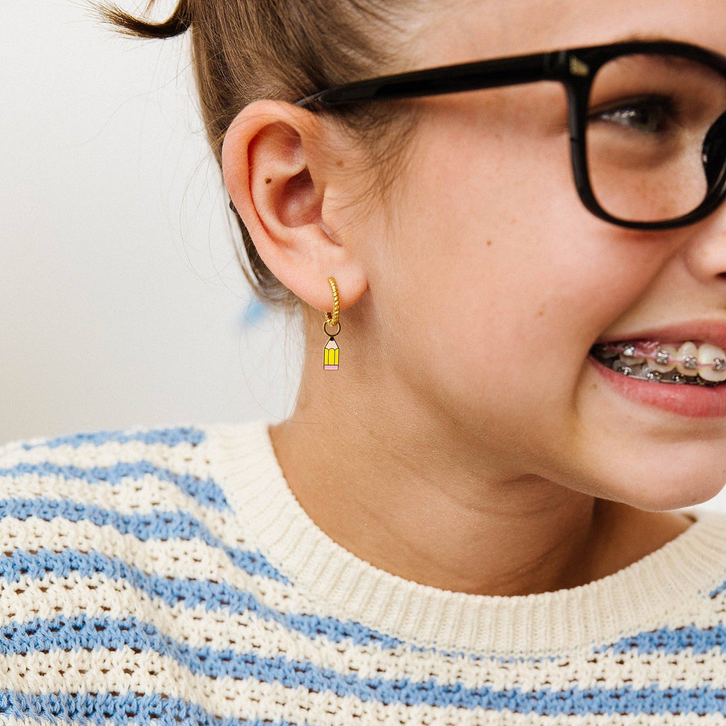 A close-up lifestyle shot of a young girl wearing a small gold huggie hoop earring with a yellow rectangular charm, paired with a blue and white striped knit sweater and black-framed glasses.