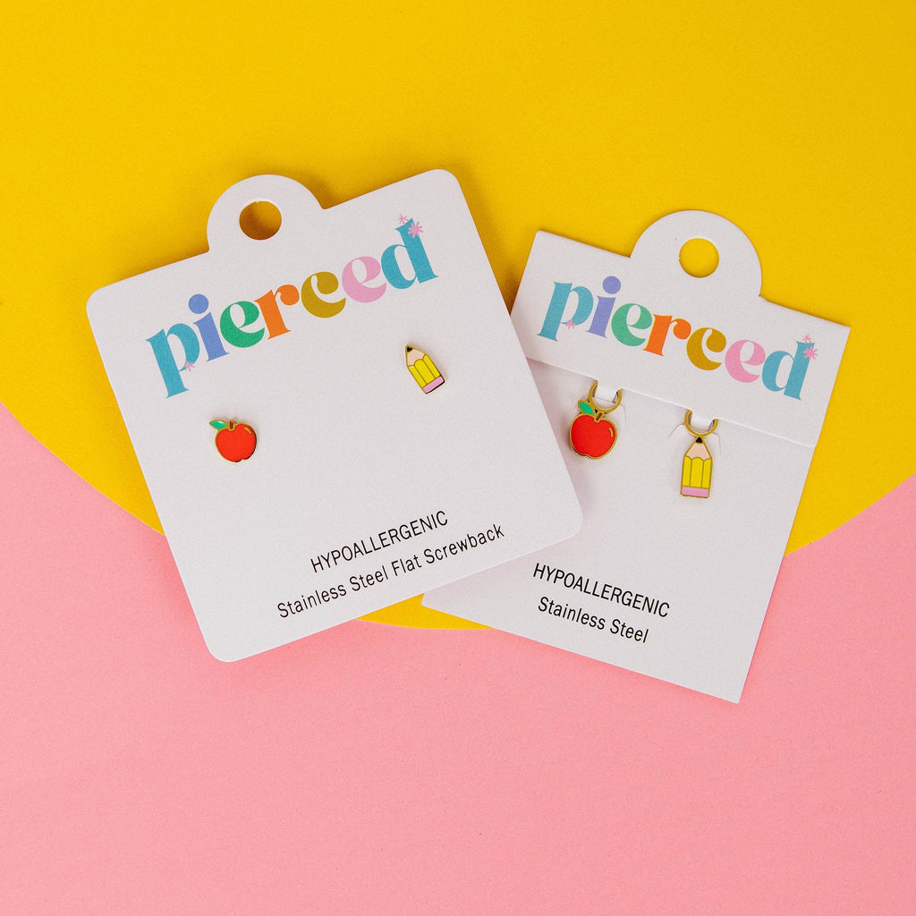Two pairs of Pierced brand stainless steel flat screwback earrings displayed on white hanging cards against a split yellow and pink background. One pair features a small red apple charm, and the other features a small yellow pencil charm.