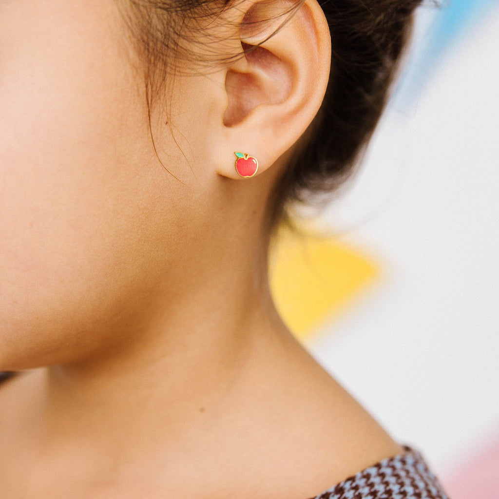 A close-up of a child's ear wearing a small apple-shaped stud earring. The earring has a red body with a green top and tiny yellow accents.