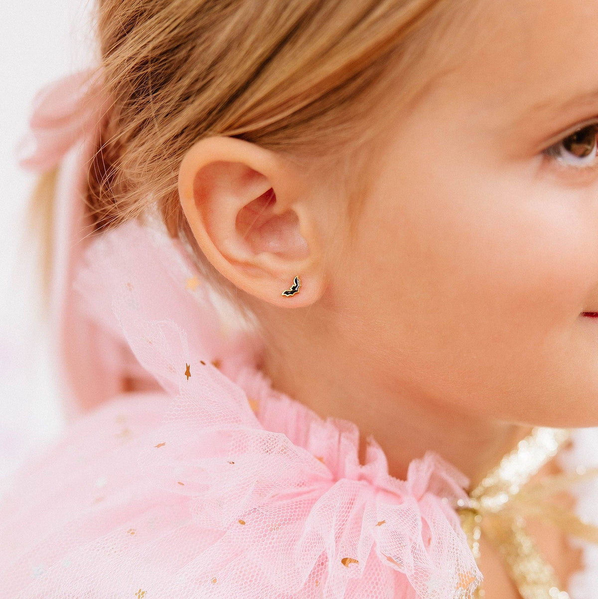 A close-up of a young child's ear wearing a small crescent moon stud earring. The child has light brown hair, wearing a pink ruffled tulle top with gold glitter details.