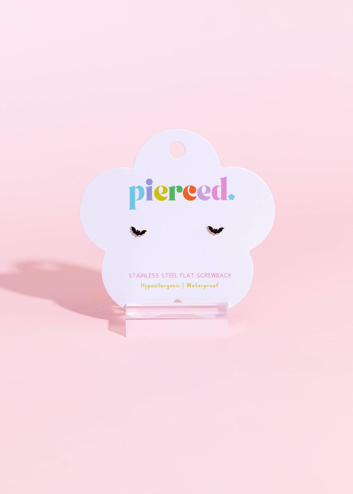 A pair of small black flatback screwback stud earrings displayed on a white cloud-shaped Pierced brand card, set against a solid pastel pink background.